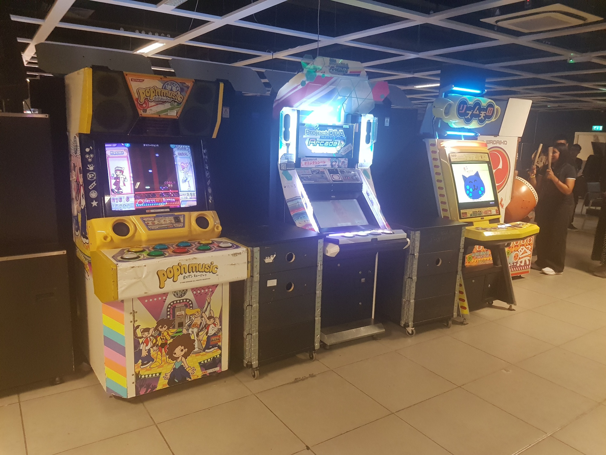 A row of rhythm games: Pop’n Music, Hatsune Miku: Project Diva, Rhythm Heaven. The side of a Taiko no Tatsujin cabinet can be seen on the right hand side