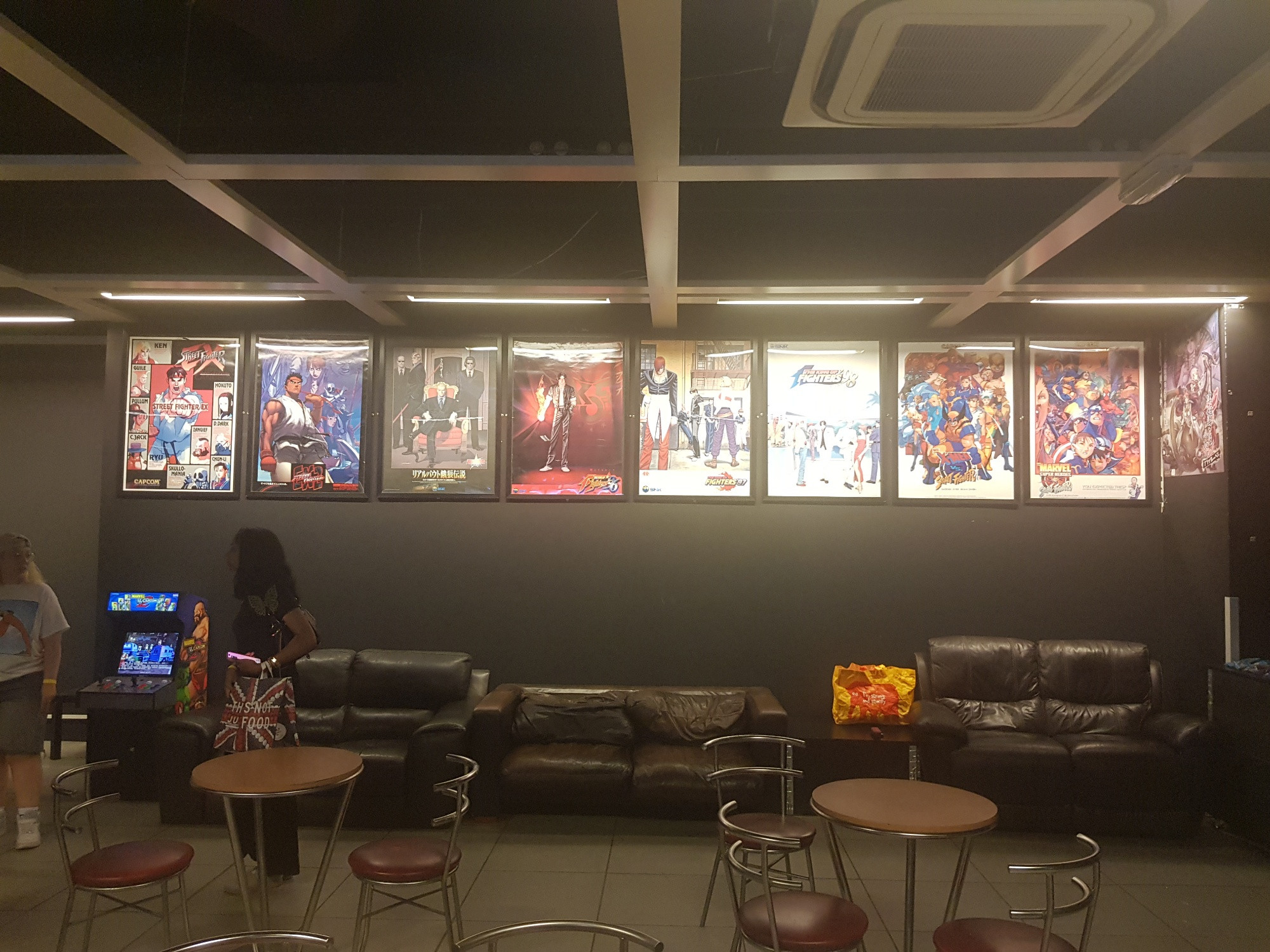 A seating area at Heart of Gaming. There are game posters (Street Fighter EX, Street Fighter EX2, Real Bout Fatal Fury, KOF96, KOF97, KOF98, X-Men vs Street Fighter, Marvel Super Heroes vs Street Fighter, Mushihimesama Futari Black Label). There’s also one of those Arcade1Up Marvel vs Capcom cabinets