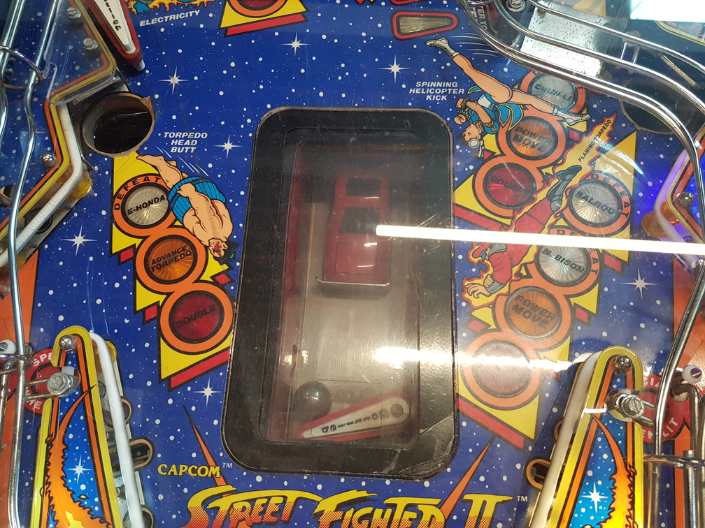 A close-up of the smash-the-car bonus stage in the Street Fighter II pinball game. Underneath a window in the playfield, there is a flipper which, at certain points in the game, can be used to flip a ball towards a toy car.